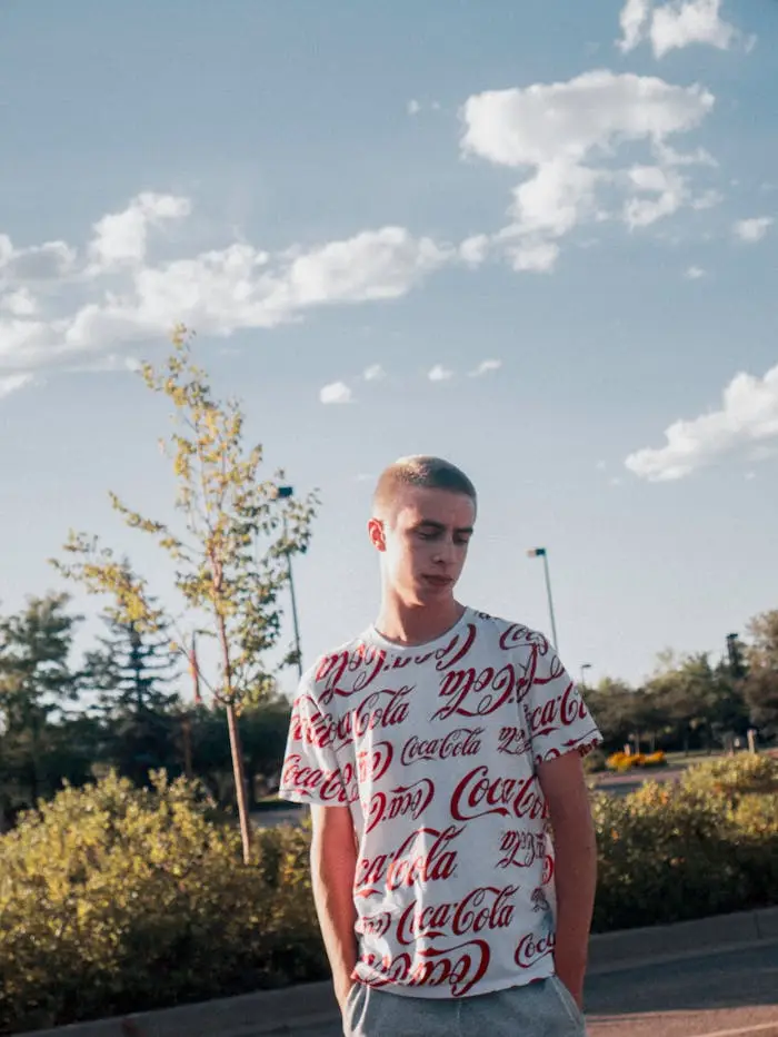 Young man in branded Coca-Cola shirt outside with sunny backdrop and vibrant greenery.
